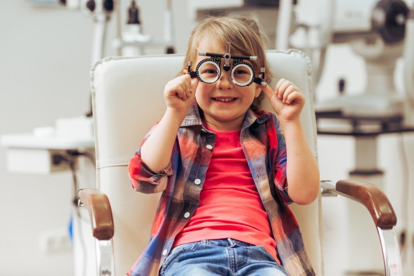 A smiling child sits on a chair in an optometrist's office holding an eye testing tool in front of her eyes.