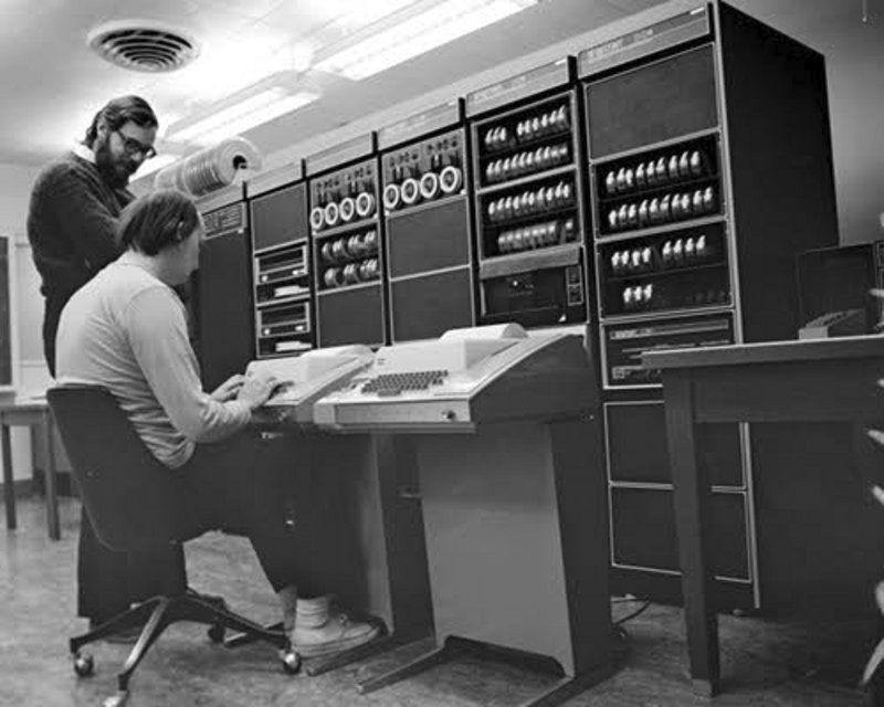 Dennis Ritchie (à gauche) discute avec Ken Thompson (deux des trois inventeurs du système Unix). Ce dernier opère sur un terminal, un équipement branché sur le GE-645, dont une partie se distingue en face d'eux.