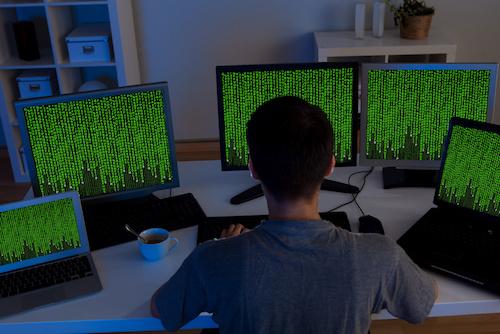 Stock image of person sitting in front of computer with multiple screens showing Matrix-like green code descending from the top of the screens to the bottom.